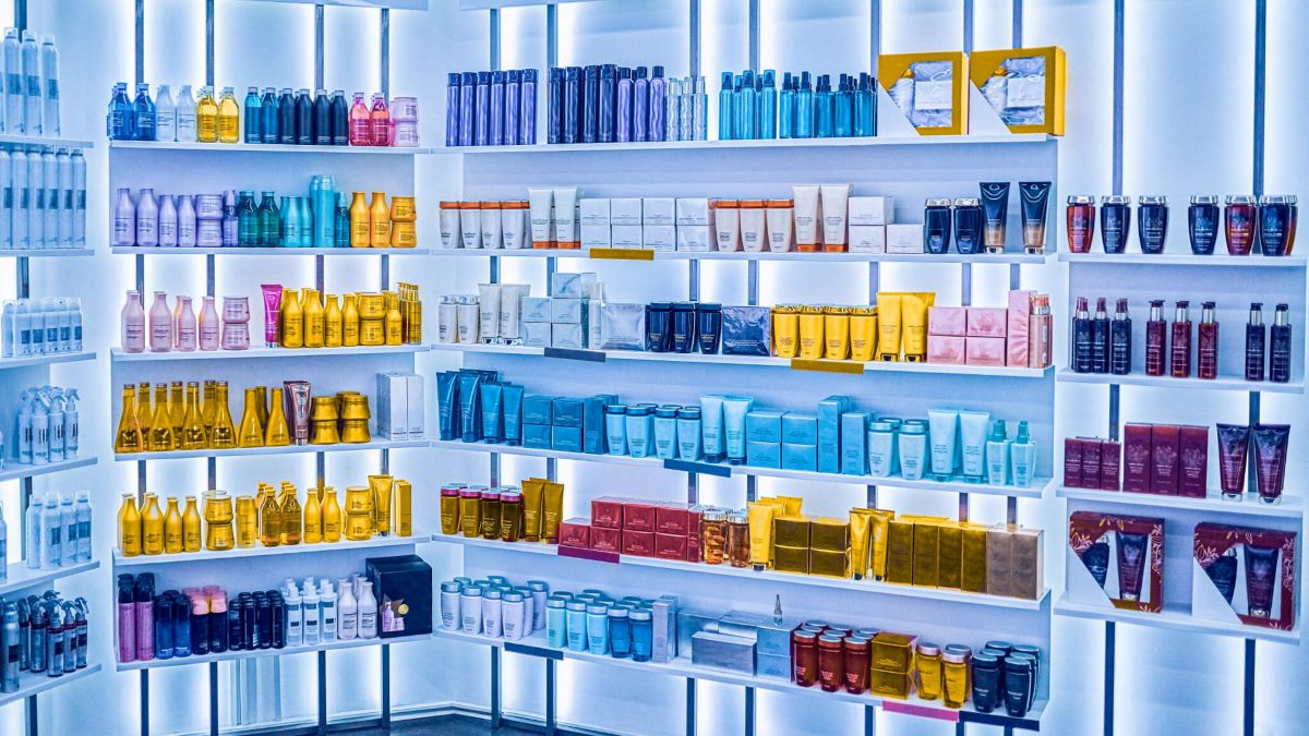A neatly organized display of various hair care and cosmetic products arranged by color on white shelves, backlit with blue-toned lighting, creating a gradient effect. The setup is clean and modern, resembling a high-end beauty supply store.