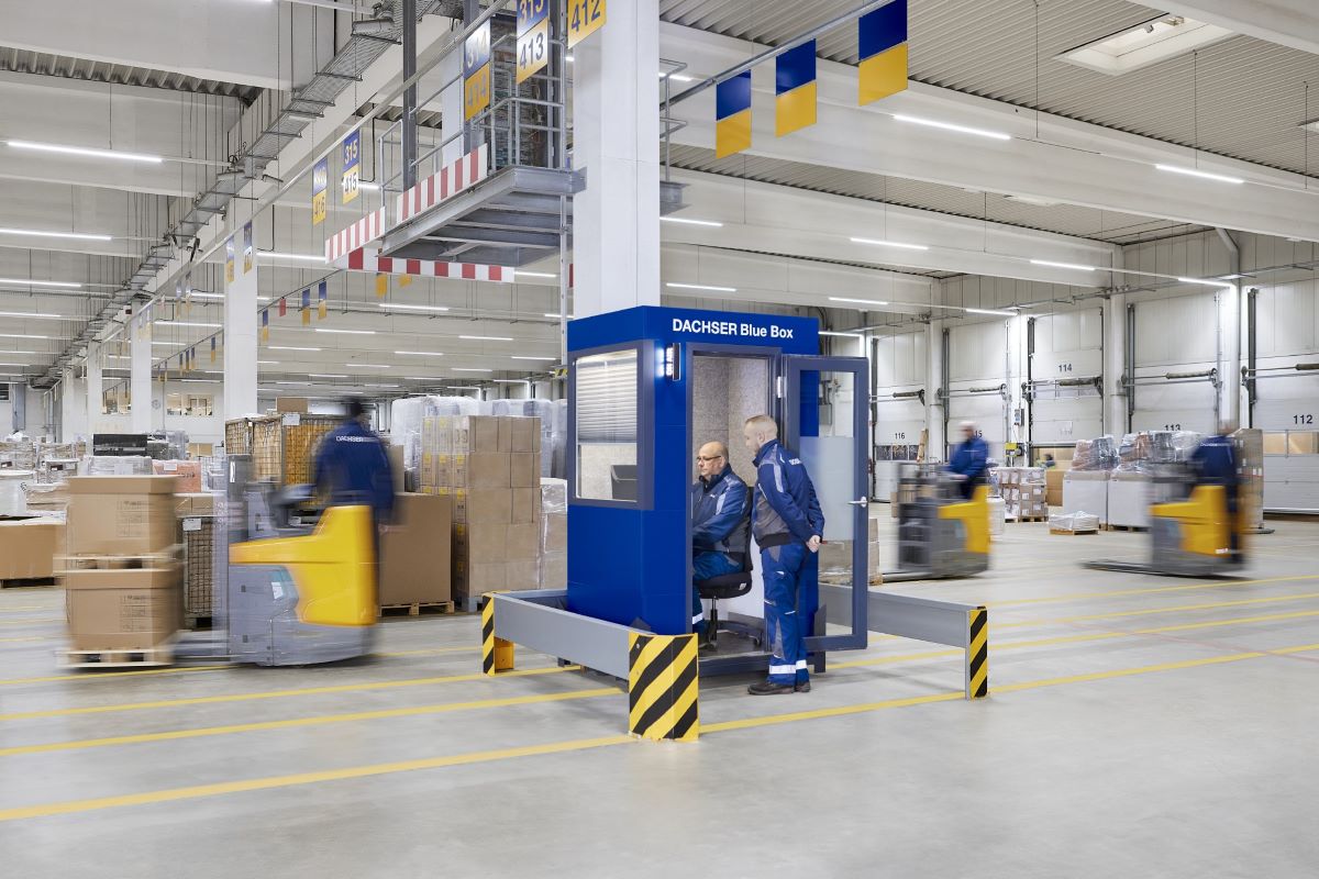 Blue Box de Dachser ubicada en un gran almacén, con dos empleados en su interior y varios montacargas en movimiento alrededor, creando un ambiente de trabajo dinámico.