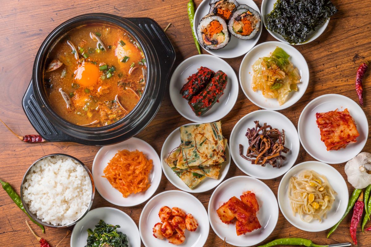 A traditional Korean meal with a hot pot of stew topped with an egg in the center, surrounded by various side dishes (banchan) including kimchi, seaweed, seasoned vegetables, pickled radish, Korean pancakes, and a bowl of steamed white rice, all arranged on a wooden table.