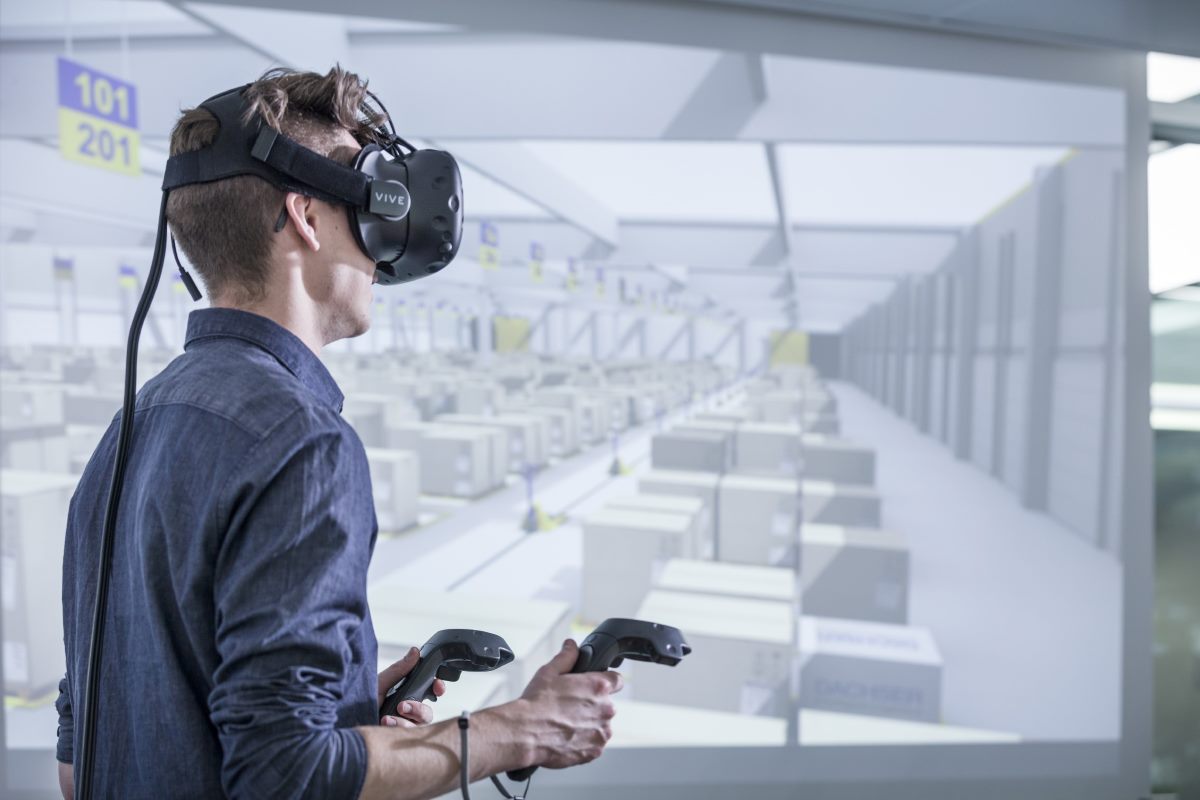A man wearing a virtual reality headset and holding controllers explores a simulated warehouse environment displayed on a large screen in the background.