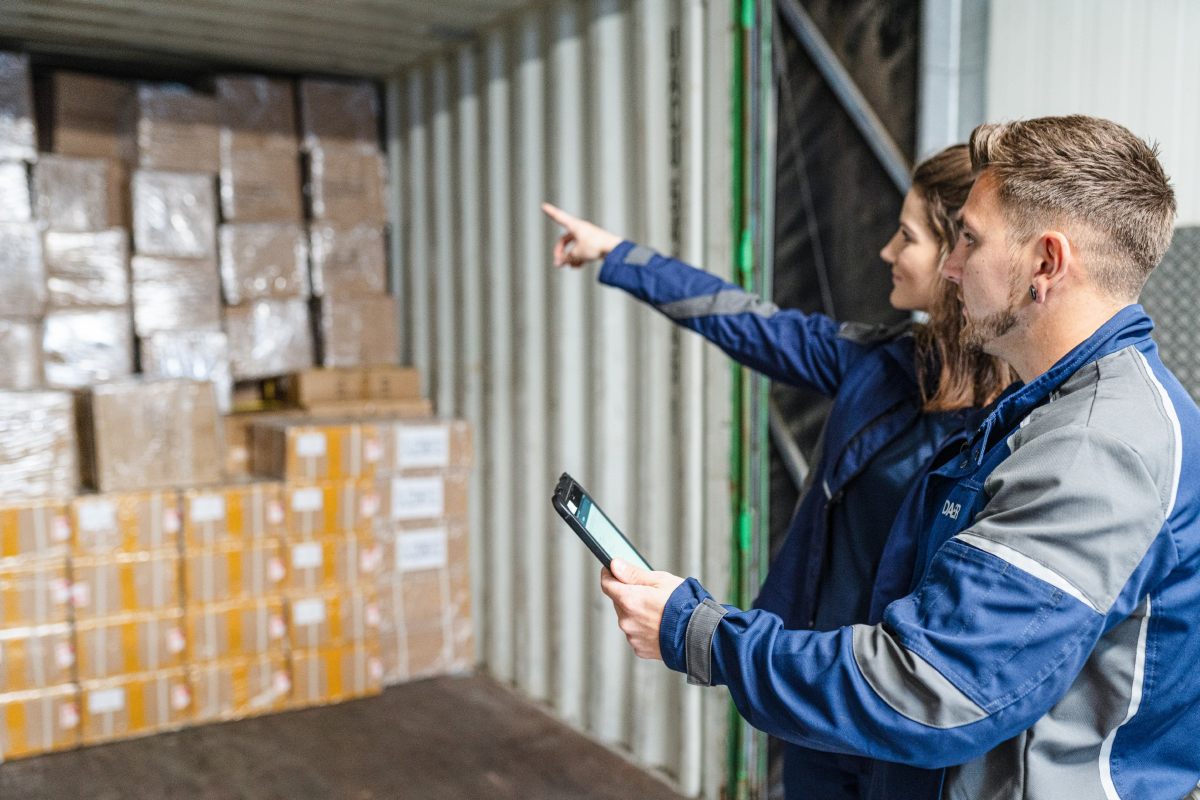 Two warehouse workers inspect and discuss stacked boxes inside a shipping container. One holds a handheld device while the other points toward the cargo.