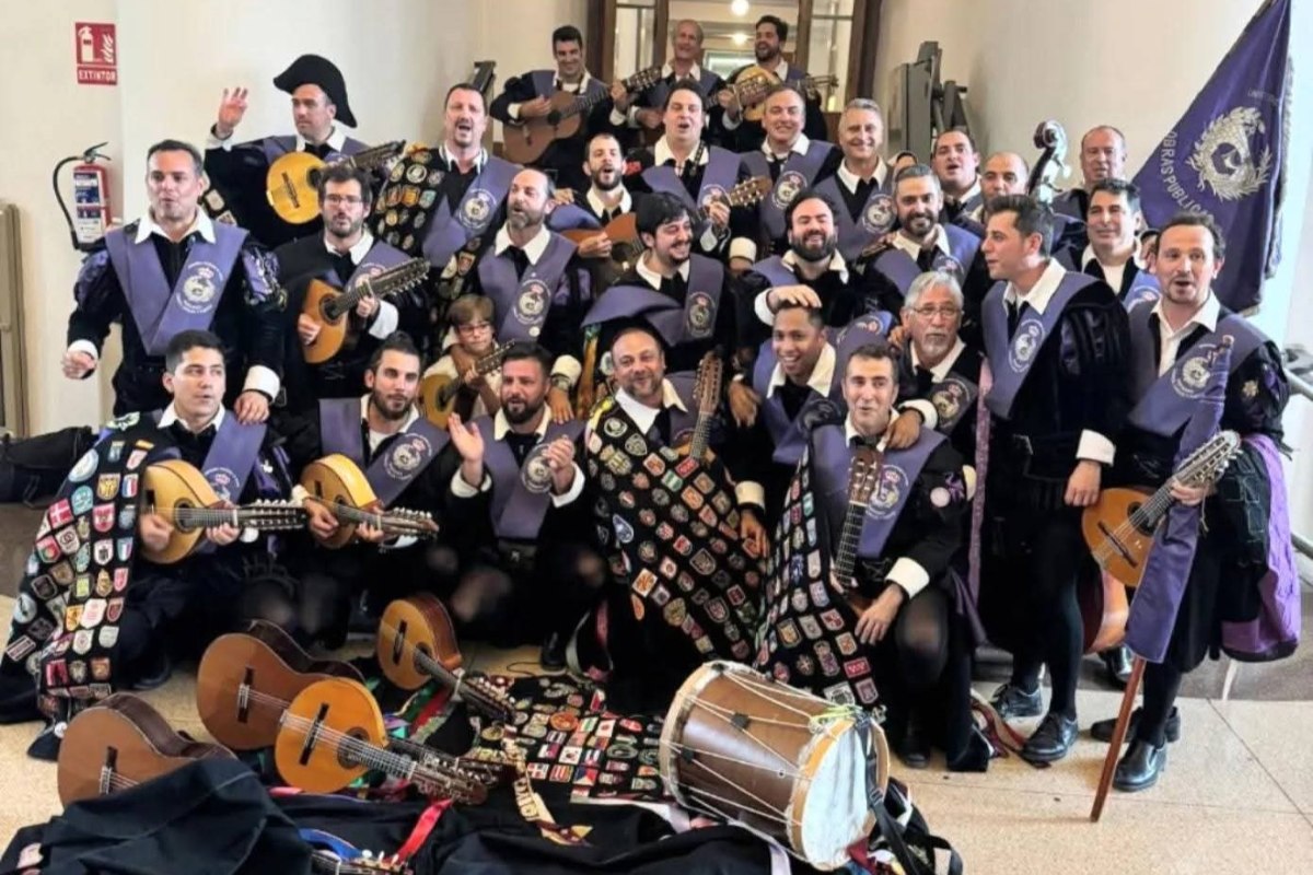 Large university tuna group posing in a hallway, wearing black capes covered with badges and purple sashes, with guitars, lutes and a drum held by the musicians and lying on the floor.
