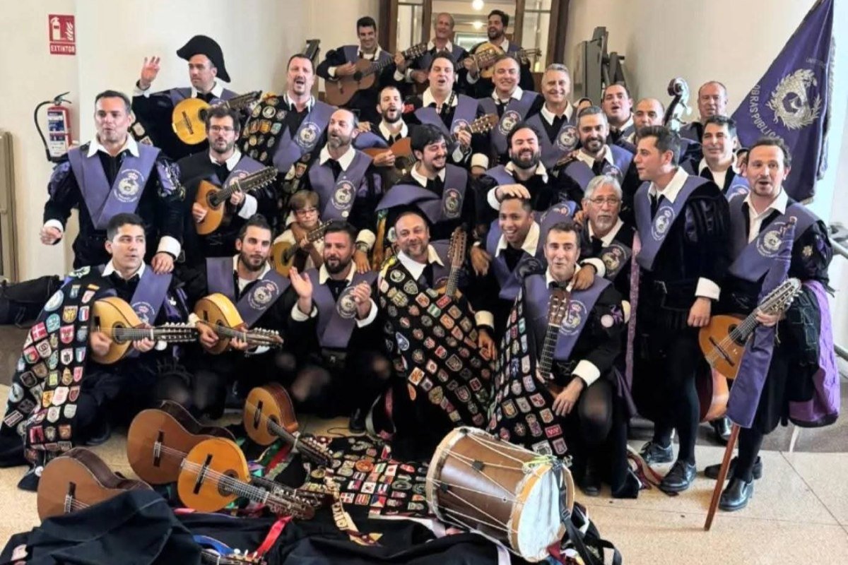 Large university tuna group posing in a hallway, wearing black capes covered with badges and purple sashes, with guitars, lutes and a drum held by the musicians and lying on the floor.