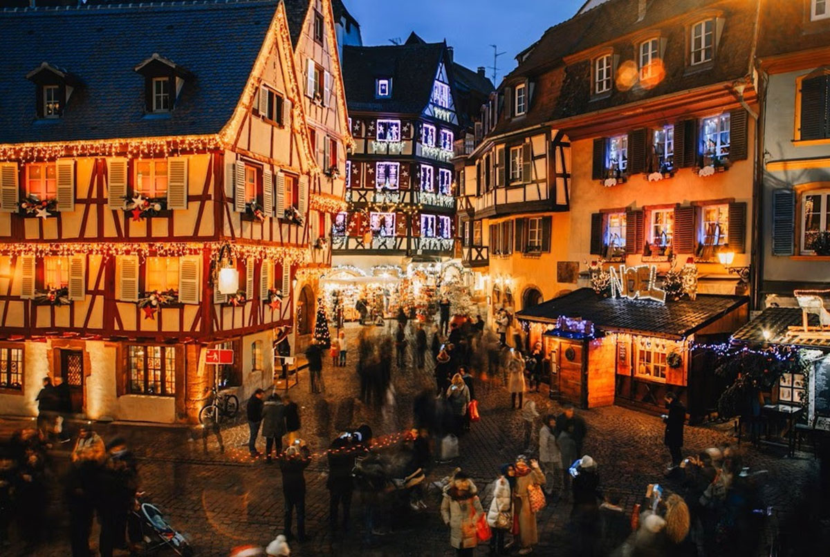 Plaza de un pueblo europeo al anochecer con casas de entramado de madera iluminadas y un mercado navideño lleno de gente.