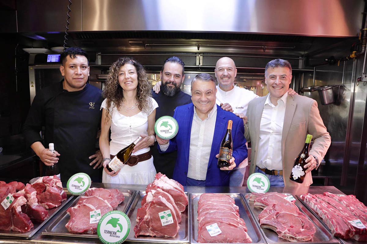 Six people pose in a professional kitchen behind trays of beef cuts, holding bottles of wine and promotional material for Asturian Veal at a gastronomic event.