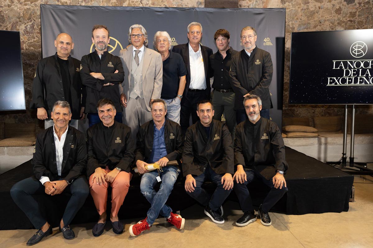 A group of chefs and representatives from Bacalao Barquero posing during a gastronomic event. Several people are wearing black jackets featuring the event's logo, and a screen displaying the text "La Noche de la Excelencia" (The Night of Excellence) appears in the background.
