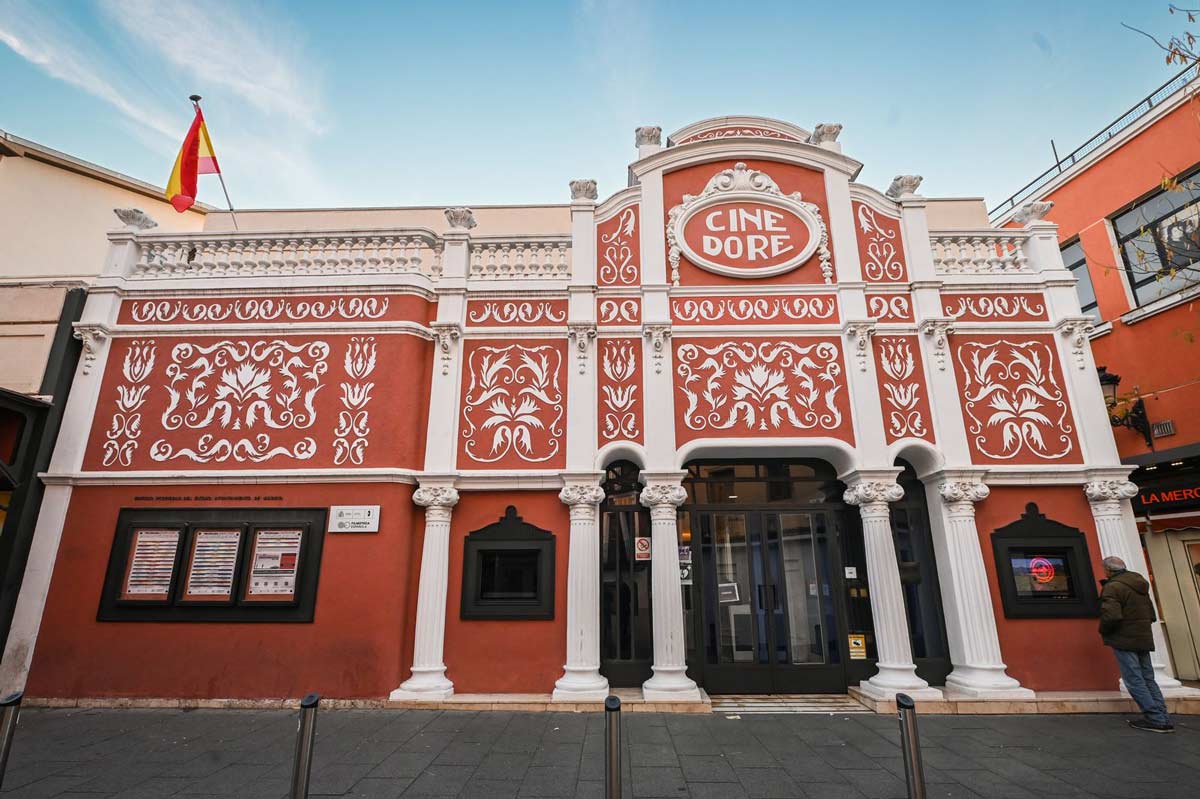 Facade of the Cine Doré, home of the Filmoteca Española in Madrid. The modernista building has a terracotta-red front decorated with raised white floral motifs, fluted columns and an upper balustrade. Above the entrance, an oval medallion bears the sign "CINE DORÉ". Display cases with the programme sit to the left, and a Spanish flag flies from the roof.