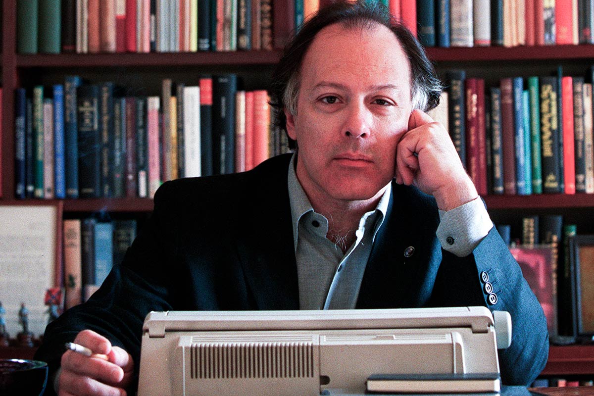 Retrato del escritor Javier Marías sentado ante una máquina de escribir, con la mano apoyada en la mejilla y un cigarrillo entre los dedos, frente a una estantería llena de libros.