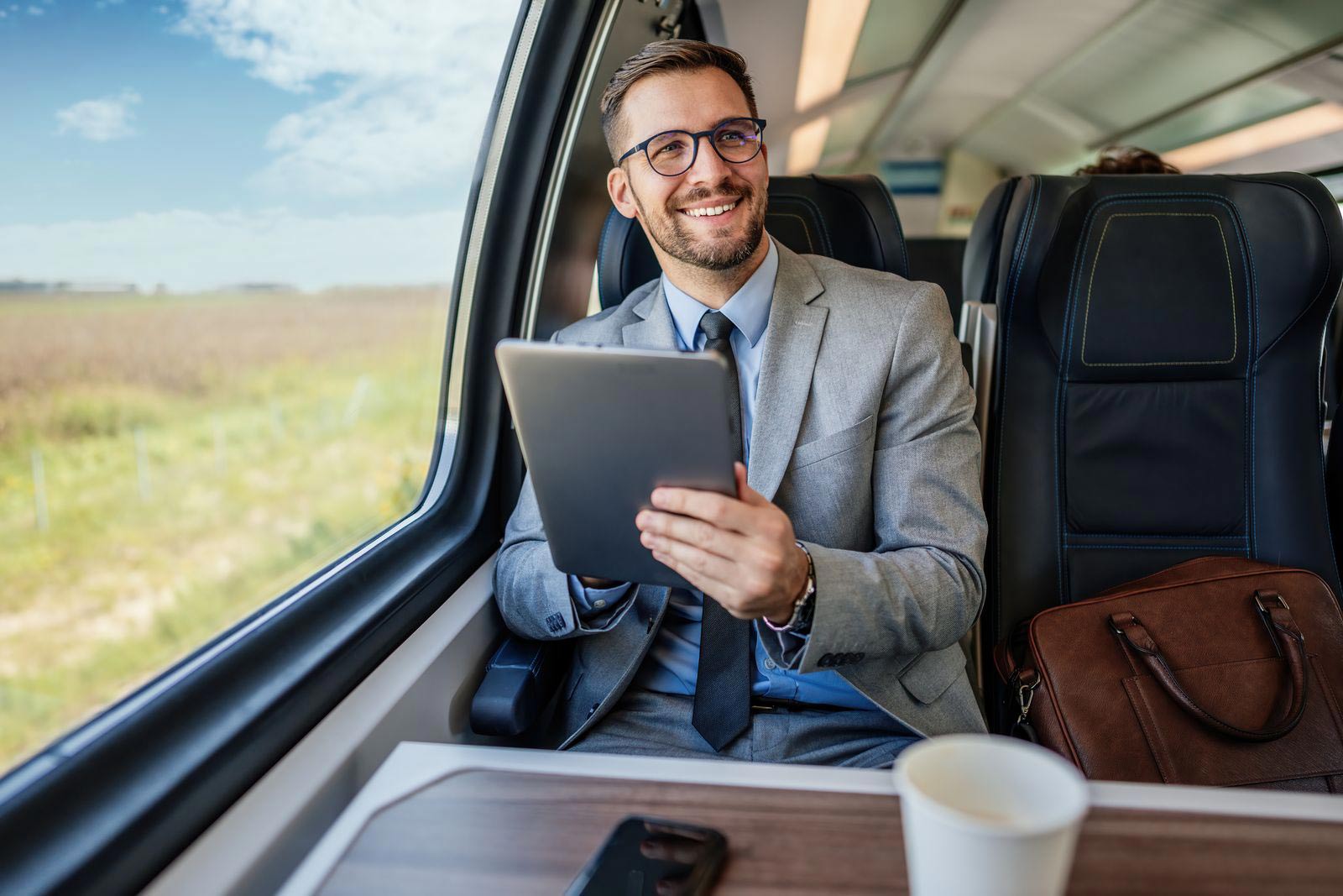 A bearded businessman in a grey suit and glasses sits by a train window, using a tablet and smiling; a coffee cup sits on the table and a leather briefcase beside him.