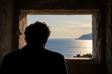 Silueta de un hombre de espaldas asomado a una ventana de piedra que enmarca el mar en calma al atardecer, con un cabo recortándose a la derecha y el sol reflejándose sobre el agua.