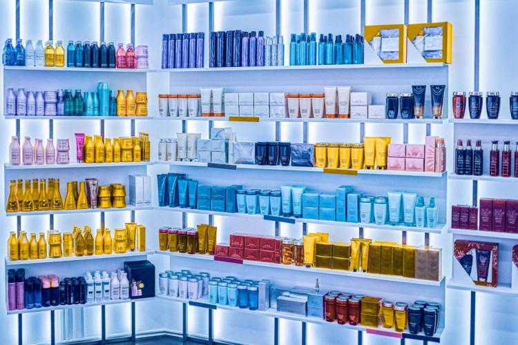 Una exhibición ordenada de diversos productos para el cuidado del cabello y cosméticos organizados por color en estantes blancos, iluminados con luz azul, creando un efecto degradado. La configuración es limpia y moderna, similar a una tienda de belleza de alta gama.