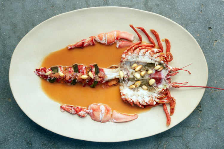 Gourmet dish of lobster cut in half and presented on an oval tray. The lobster is decorated with peanuts and seaweed, and surrounded by sauce.