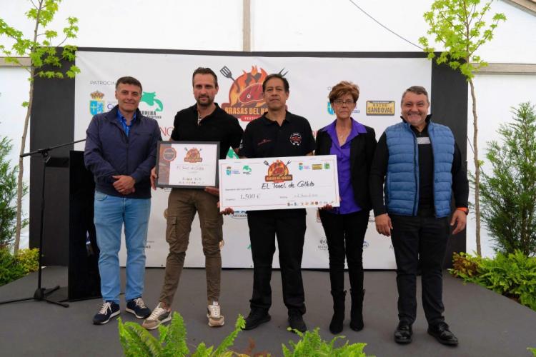 A group of five people from Ronel de Colloto restaurant standing on a stage, posing for a photo. The person in the center is holding a certificate and a large check for 1,500 euros. The background features a banner with logos and the text Brasas del Narcea Festival.