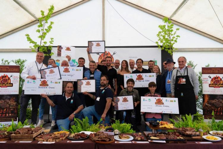 Prize winners of the first edition of the Best Barbecue Contest in Spain with Asturian Beef. A large group of people posing on a stage, each holding certificates and large checks. In the foreground, there is a table displaying various grilled meats and dishes. The background features banners with logos and the text Brasas del Narcea Festival.