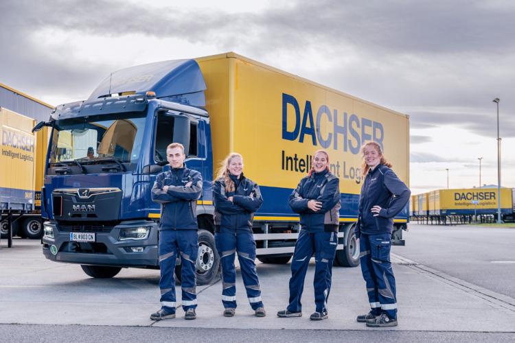 A group of four DACHSER employees stands confidently in front of a DACHSER branded truck. The truck has a blue cab and a yellow trailer with the company's logo and slogan "Intelligent Logistics" clearly visible. The employees are dressed in company uniforms, consisting of blue jackets and pants with reflective stripes. The background features additional DACHSER trailers and a partly cloudy sky.