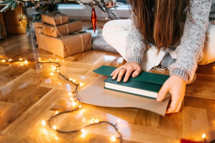 A person wrapping a green book in brown paper while sitting on a wooden floor surrounded by glowing Christmas lights and gift boxes under a decorated tree.