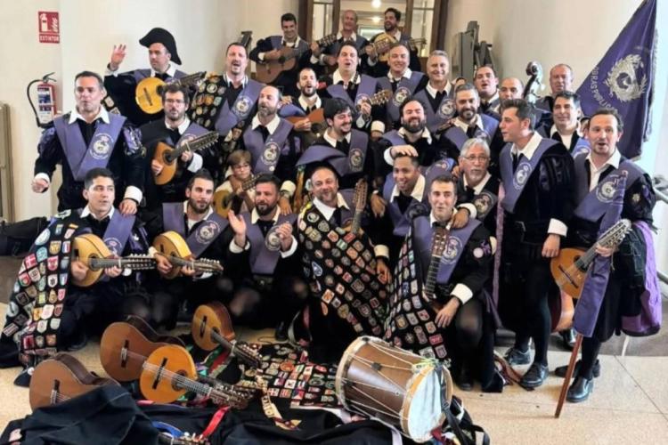 Large university tuna group posing in a hallway, wearing black capes covered with badges and purple sashes, with guitars, lutes and a drum held by the musicians and lying on the floor.