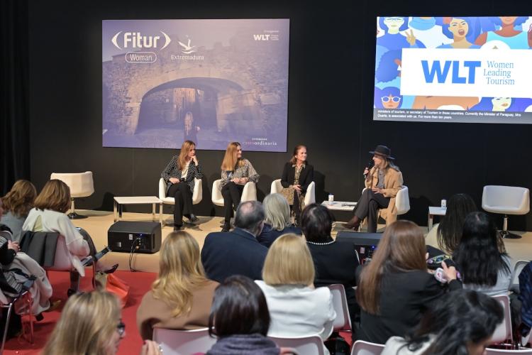 Panel en FITUR Woman (Women Leading Tourism): cuatro mujeres sentadas en el escenario, una hablando con micrófono, con público en primer plano y pantallas con los logos FITUR Woman y WLT.
