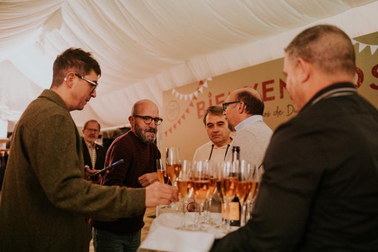 In a tent-like space with a white fabric roof and decorative pennants, several people chat around a high table. On the table are glasses filled with sparkling/rosé wine and bottles, while in the background a large welcome sign reinforces the festive and celebratory atmosphere.
