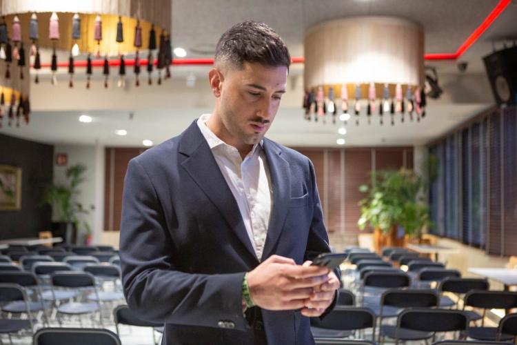 Juan Pedro Martinez in a dark suit checking his phone in an empty conference room with folding chairs and hanging lamps.