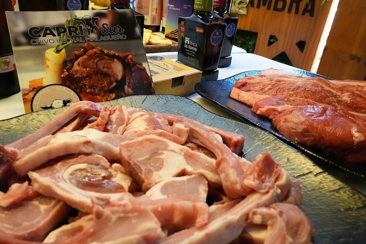 Close-up display of raw goat cuts and other meats arranged on serving platters at a food promotion event, with olive oil, wine bottles, and a sign promoting Málaga milk-fed goat in the background.