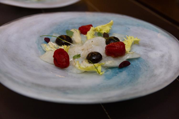 Sliced cod served on a white and blue plate, garnished with red tomatoes, black olives, lettuce leaves, and small decorative sprouts.