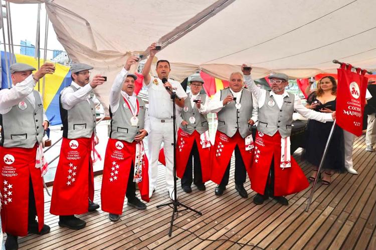 A group of people are raising glasses of red wine for a toast under a tent set up on the deck of the Juan Sebastián Elcano. They are wearing the red aprons and gray vests of the Cofradía Gastronómica del Cocido Madrileño (Madrid Stew Gastronomic Brotherhood), while a person in a white uniform speaks into a microphone. The brotherhood's banner is visible on the right.