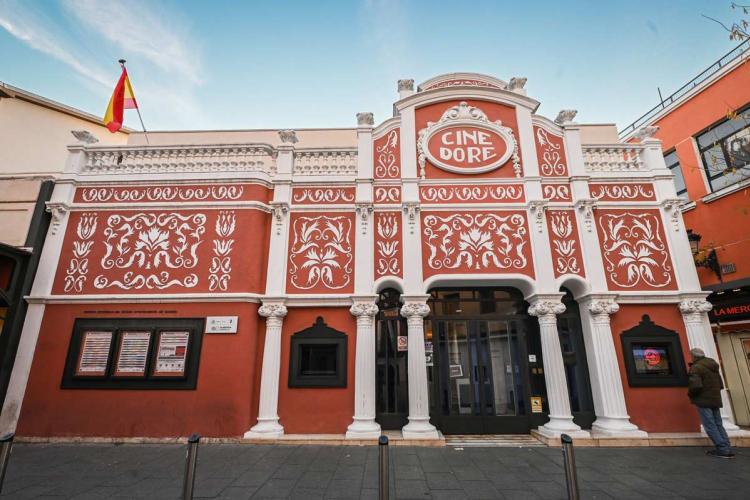 Facade of the Cine Doré, home of the Filmoteca Española in Madrid. The modernista building has a terracotta-red front decorated with raised white floral motifs, fluted columns and an upper balustrade. Above the entrance, an oval medallion bears the sign "CINE DORÉ". Display cases with the programme sit to the left, and a Spanish flag flies from the roof.