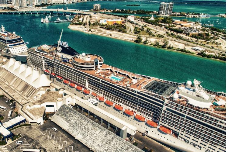 Aerial view of a large cruise ship docked at a port, with the city and bay in the background.