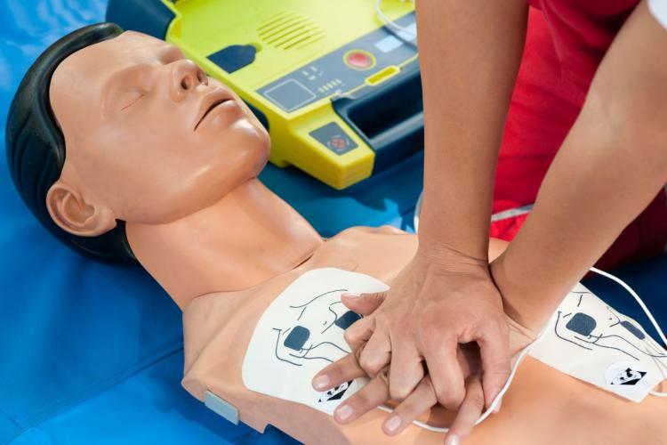 Person performing cardiopulmonary resuscitation (CPR) chest compressions on a training manikin with automated external defibrillator (AED) pads attached to the chest. An open AED is positioned beside the manikin during a first aid training session.