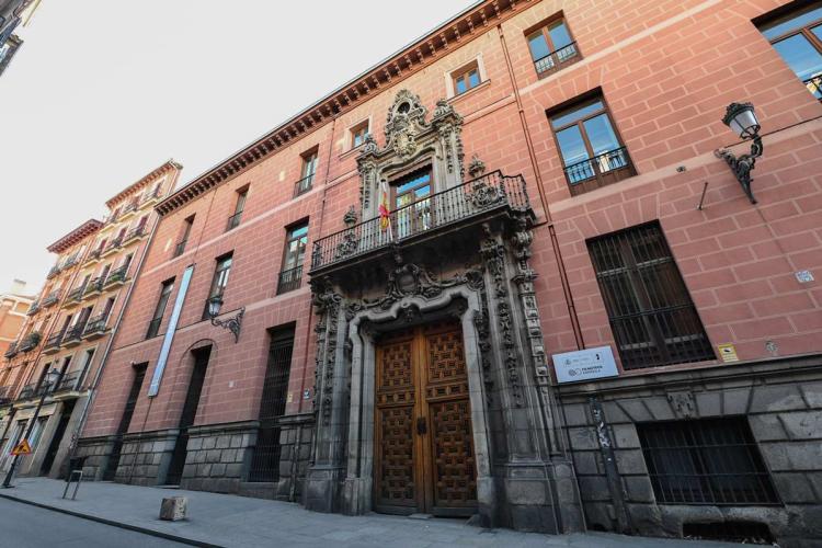 Facade of the Palacio del Marqués de Perales, home of the Filmoteca Española in Madrid, seen at an angle from the street. The salmon-pink wall rises from a granite base and opens into a carved stone Baroque doorway with a heraldic coat of arms and a double wooden door. Above it, a wrought-iron balcony flies a flag. The Filmoteca Española sign appears to the right.