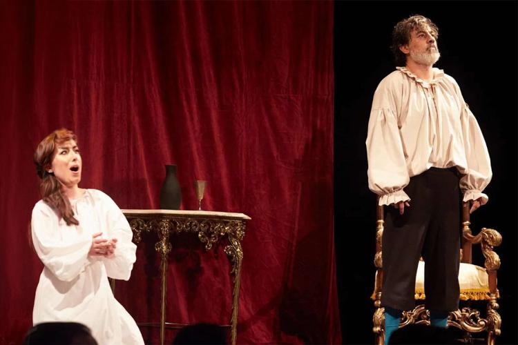 Escena de zarzuela: una soprano vestida de blanco canta arrodillada junto a una mesa dorada mientras el barítono Marco Moncloa, en traje de época, permanece de pie sobre un sillón ante un cortinón rojo
