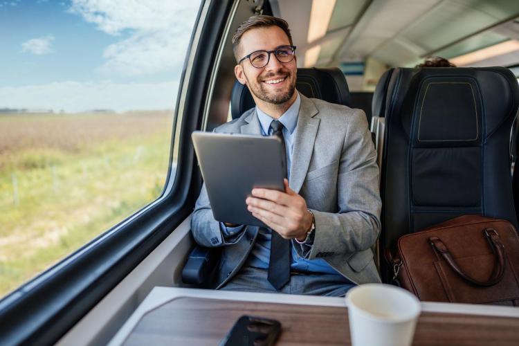 A bearded businessman in a grey suit and glasses sits by a train window, using a tablet and smiling; a coffee cup sits on the table and a leather briefcase beside him.