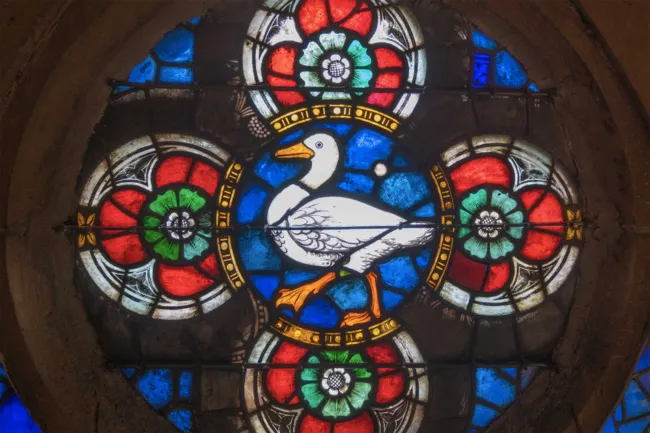 Circular stained glass window with a white goose in the center surrounded by red, green and blue floral motifs.