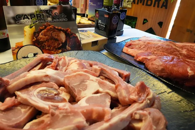 Close-up display of raw goat cuts and other meats arranged on serving platters at a food promotion event, with olive oil, wine bottles, and a sign promoting Málaga milk-fed goat in the background.