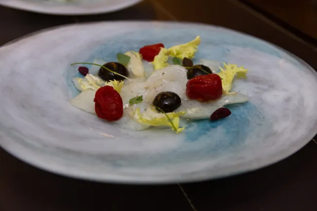 Sliced cod served on a white and blue plate, garnished with red tomatoes, black olives, lettuce leaves, and small decorative sprouts.