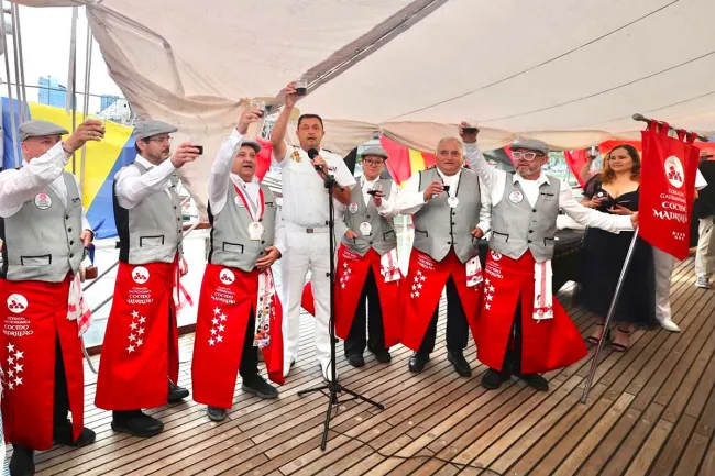 A group of people are raising glasses of red wine for a toast under a tent set up on the deck of the Juan Sebastián Elcano. They are wearing the red aprons and gray vests of the Cofradía Gastronómica del Cocido Madrileño (Madrid Stew Gastronomic Brotherhood), while a person in a white uniform speaks into a microphone. The brotherhood's banner is visible on the right.