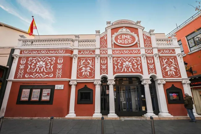 Facade of the Cine Doré, home of the Filmoteca Española in Madrid. The modernista building has a terracotta-red front decorated with raised white floral motifs, fluted columns and an upper balustrade. Above the entrance, an oval medallion bears the sign "CINE DORÉ". Display cases with the programme sit to the left, and a Spanish flag flies from the roof.