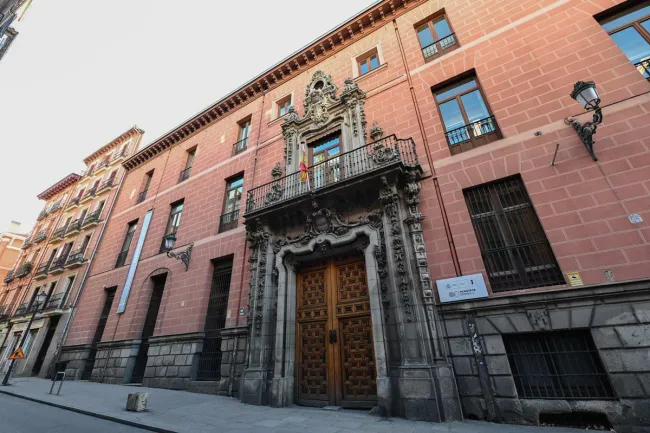 Facade of the Palacio del Marqués de Perales, home of the Filmoteca Española in Madrid, seen at an angle from the street. The salmon-pink wall rises from a granite base and opens into a carved stone Baroque doorway with a heraldic coat of arms and a double wooden door. Above it, a wrought-iron balcony flies a flag. The Filmoteca Española sign appears to the right.