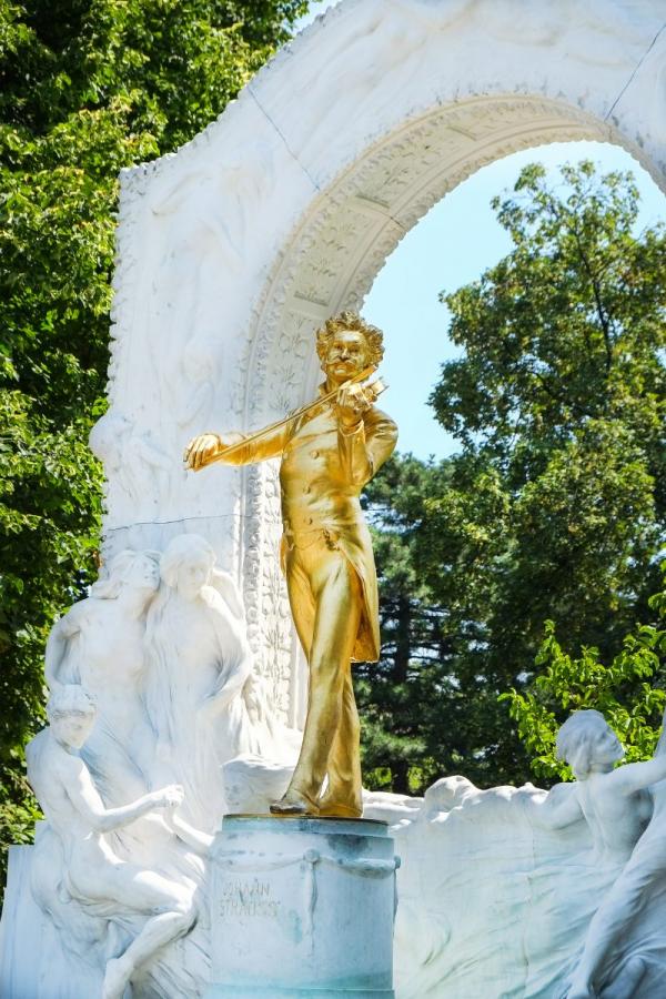 Full view of the Johann Strauss II monument in Vienna, featuring a golden statue surrounded by a white marble arch with ornate sculptures, set in a green park.