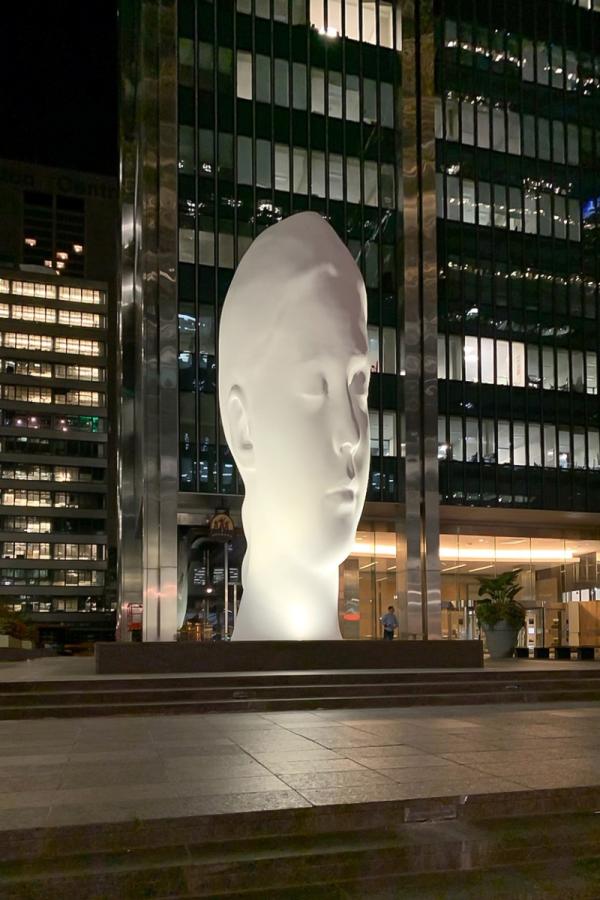 Dreaming, by Jaume Plensa, large illuminated sculpture of a serene white head at night in front of a modern glass building.