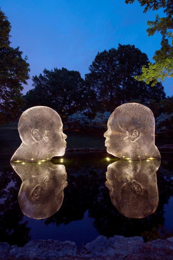 Awilda and Irma, by Jaume Plensa, two large mesh head sculptures facing each other, glowing at night and reflecting on the water with trees in the background.