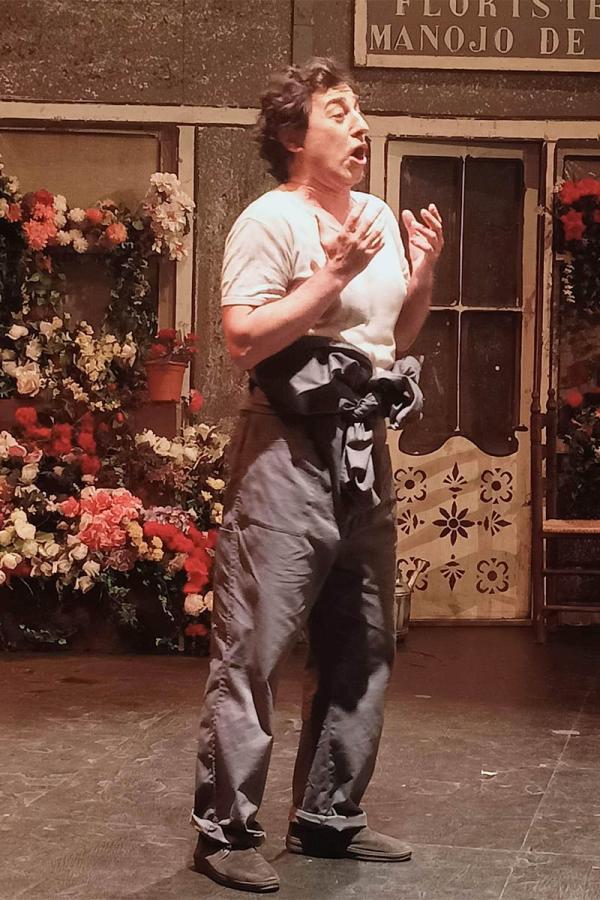 Baritone Marco Moncloa sings with his hands raised in front of a florist’s shop set, in a performance of La del manojo de rosas