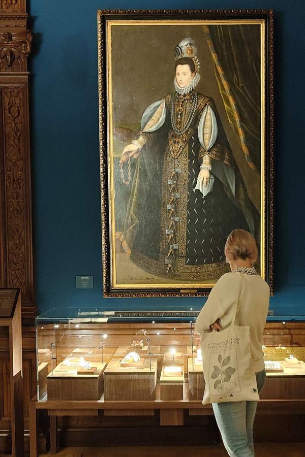 A visitor looks at a large full-length portrait of a 16th-century noblewoman in an embroidered black gown, hung on a blue gallery wall above a lit jewellery display case