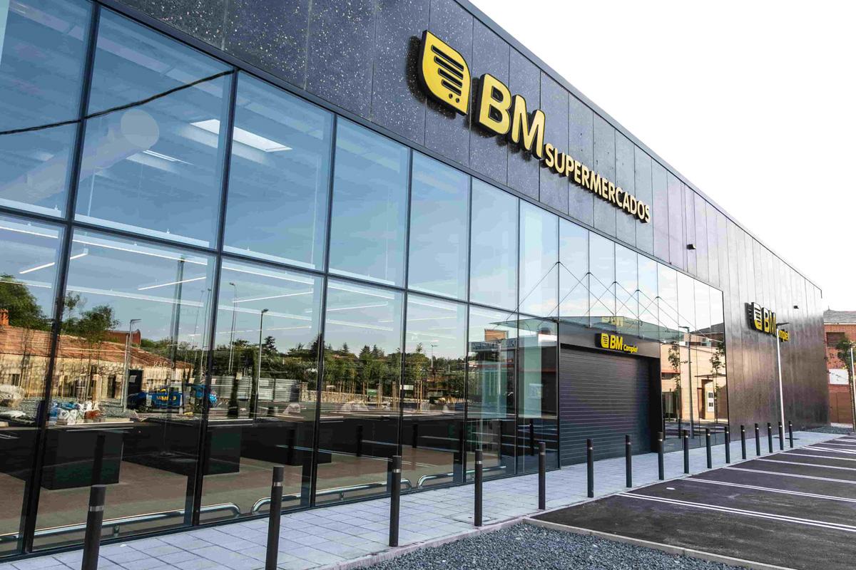 Facade of a BM supermarket with a modern design. The building features large glass windows reflecting the exterior, including trees and nearby buildings. The BM Supermercados logo, in yellow and white letters, is prominently displayed at the top of the facade. The parking area in front of the supermarket is empty.